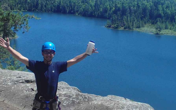 A person wearing safety gear stands on a rock overlook above a blue body of water. They have their arms spread wide in the air, and are holding a water bottle in one hand. 